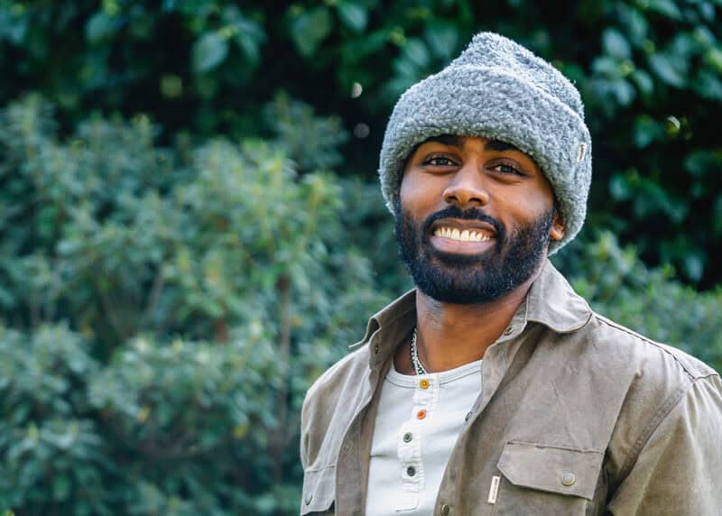 Man in beanie and coat stands outside in front of large green trees